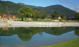 A quiet park with a clear pond and green meadows. In the background, mountains and playground equipment can be seen.