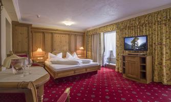 A cozy hotel room with wooden details and red carpets. There is a large bed, a television, and a balcony with daylight.