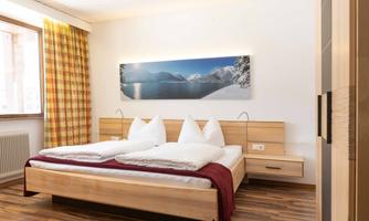 A cozy bedroom with a large bed and wooden paneling. On the wall hangs a picture of a snow-covered landscape, and the windows have curtains that match the wood decor.