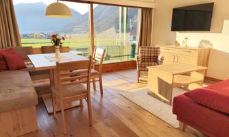 A bright and modern living room with large windows and a view of the mountains. There is a dining table, chairs, and a cozy sofa.