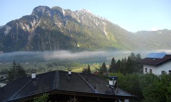 A picturesque mountain panorama with high peaks and gentle wisps of fog. In the foreground, cozy houses and lush trees can be seen.