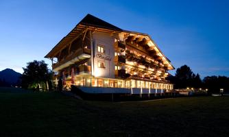 A beautiful hotel building at dusk, illuminated with warm light. In the background, gentle hills and trees are visible.