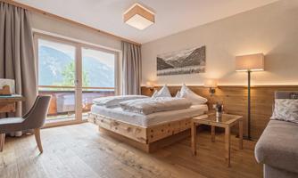 A bright hotel room with a large bed and wooden floor. Large windows offer a view of the mountains.