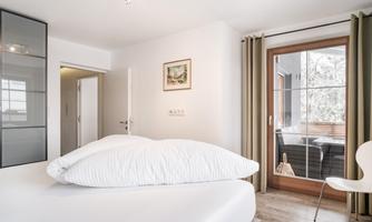 A bright, modern bedroom with a large bed and a cozy armchair. The windows are open and provide access to a balcony.