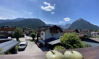 A picturesque mountain landscape with houses and a green garden. In the foreground, stylish green apples are placed on a wooden table.