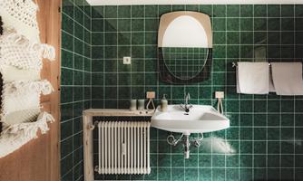 A modern bathroom with green tiles and a white sink. A round mirror and towels are also visible.