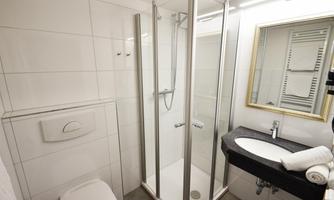 A modern bathroom with a shower, sink, and mirror. The walls are covered in light tiles and there are fresh towels.