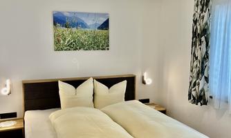 A modern bedroom with a large bed and two pillows. A picture of a landscape hangs on the wall, and there are bright curtains and pleasant lighting.