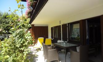 A terrace with a dining table and chairs, surrounded by plants. In the background, windows and a sunny atmosphere are visible.