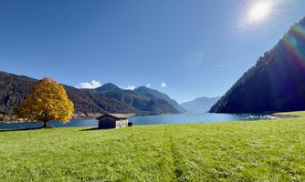 A picturesque landscape with a clear lake and snow-capped mountains in the background. A solitary tree and a small cabin stand on the green meadow.
