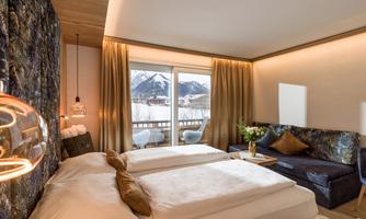 A modern hotel room with two single beds and a cozy seating area. Large windows offer a view of the mountains.