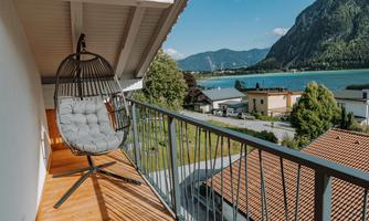 A comfortable hanging chair on a balcony with a view of the lake and the mountains. The surroundings are green and inviting.