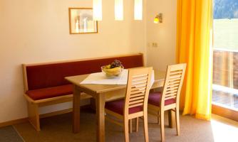 A bright dining area with a wooden table and chairs. Large windows with yellow curtains let in plenty of light.