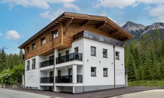 A modern building in alpine style with wooden cladding and balconies. It is located in a quiet environment, surrounded by trees and mountains.