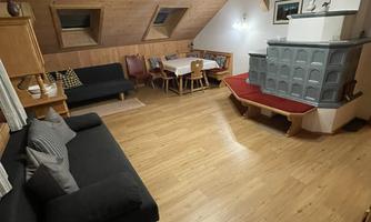 A cozy living room with a wooden ceiling and wooden floor. It features a seating area, a dining table, and a heating stove.