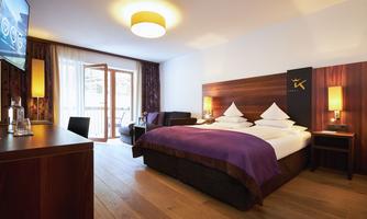 A modern hotel room with a large double bed and a cozy seating area. The large window front lets in plenty of light and provides access to a balcony.