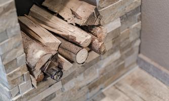 A pile of wood lies in an open fire pit. The surroundings consist of stones and a wooden floor.