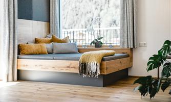 A cozy window bed with colorful cushions and a blanket. The room has large windows and a view of nature.