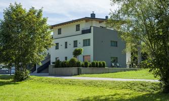 A modern building surrounded by green spaces and trees. The sky is clear and the landscape is inviting.