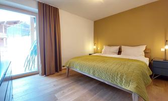 A modern bedroom with a double bed and a green bed cover. Large windows provide plenty of daylight and offer a view outside.