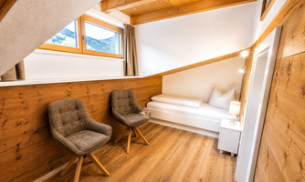 A cozy room with wooden-paneled walls and a sloping roof window. There are two chairs and a bed with white pillows.