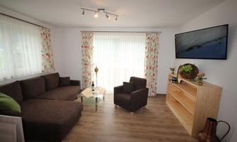 A cozy living room with a large sofa and two armchairs.  
A lot of light comes through the windows, and a television as well as a wooden shelf are present in the room.