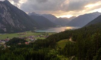 A picturesque mountain landscape with a lake in the foreground. The sun is setting behind the mountains, casting a warm light over the scene.