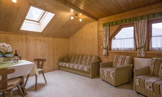 A cozy living space with a wooden ceiling and windows. The room features a table, chairs, and two comfortable sofas.