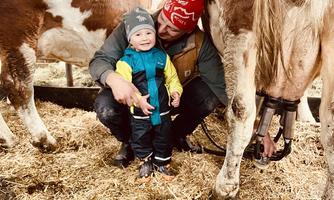 A man kneels next to a small child in a barn. They are standing among cows and straw.