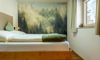A cozy bedroom with a wooden bed and a soothing forest wallpaper. The window lets in soft light.