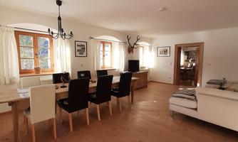 A bright dining room with hardwood floors and a large table. The walls are decorated with pictures, and there is a cozy living area.