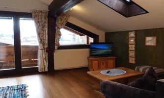 A cozy living room with wooden flooring and large windows. There is a sofa, a table, and a television, as well as decorative elements on the wall.