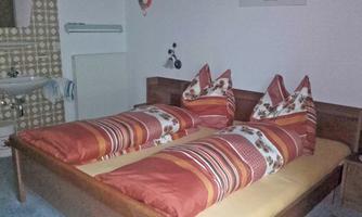 A cozy bedroom with a double bed and colorful cushions. In the background, a sink and a tiled wall are visible.