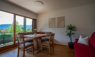 A cozy living area with a wooden table and chairs. Large windows offer a view of the green landscape outside.