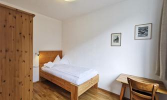 A simple bedroom with a wooden bed and a desk. The walls are white and there are a few pictures on the wall.