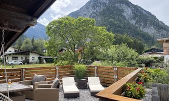 A cozy terrace with modern furniture and plants. In the background, a majestic mountain rises under a partly cloudy sky.