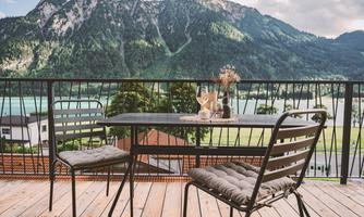 A beautiful balcony with a table and two chairs, surrounded by a picturesque mountain landscape. In the background, the clear lake and the green trees can be seen.