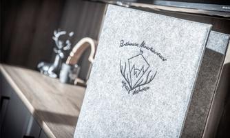 An elegant menu book with an appealing design is lying on a wooden shelf. In the background, stylish kitchen utensils are visible.