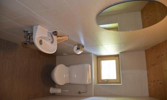 A modern bathroom with a toilet, a sink, and a window. The walls are light tiled and the ceiling is made of wood.