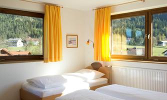 A cozy bedroom with two single beds and large windows. The yellow curtains let in a lot of light and offer a view of the surrounding nature.