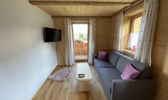 A cozy wooden living room with a gray sofa and a small table. Large windows let in plenty of light and provide access to the balcony.