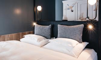 A stylish bedroom with a double bed and modern, upholstered headboards. The walls are painted in a dark shade, and the elegant lighting creates a cozy atmosphere.