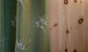A green curtain with deer motifs contrasts with a light wooden wall. The combination gives the room a natural and cozy atmosphere.