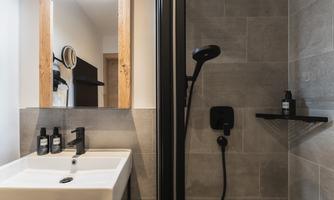 A modern bathroom with an elegant shower and sink. The walls are tiled with gray tiles, and there are toiletries on a shelf.