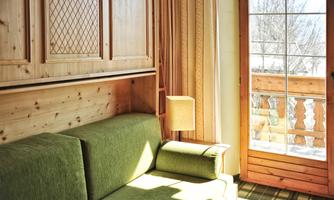 A cozy room with a green sofa and wooden furniture. Large windows offer a view of the balcony and allow plenty of light in.