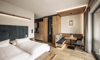 A modern room with a double bed and wooden furniture. The room features a dining area with a cozy bench and large windows.
