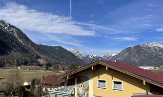 A picturesque mountain landscape with snow-covered peaks and a bright blue sky. In the foreground, the roofs of houses and green meadows are visible.