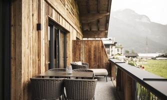 A cozy wooden balcony with a table and chairs. In the background, there are mountains and a green meadow.