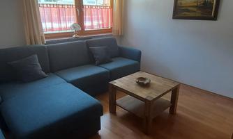 A cozy living room with a blue sofa and a wooden table. The room has large windows that let in plenty of daylight.