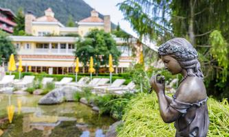 A beautiful garden with a statue of a girl and a pond. In the background, a hotel with umbrellas and trees can be seen.